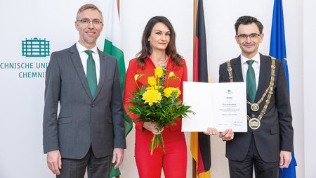 Eine Frau mittleren Alters in einem Kleid und langen Haaren hält Blumen und eine Urkunde. Links neben ihr steht ein Mann mittleren Alters mit Bart und Brille. Rechts von ihr ein Mann mittleren Alters mit Brille. Beide tragen einen Anzug.