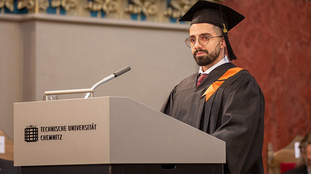 Ein junger Mann mit Brille in festlicher Kleidung steht hinter einem Rednerpult.