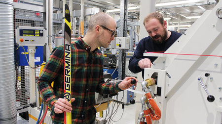 Zwei Männer arbeiten an einer Maschine, einer hält einen Ski in der Hand.