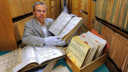 Ein älterer Mann mit Brille hält ein großes und altes Buch aufgeschlagen in die Kamera.