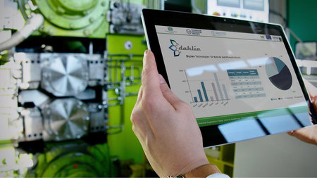 Zwei Hände halten einen Tablet-Computer, auf dem Display erscheinen Diagramme und das Logo des Projektes dahlia.