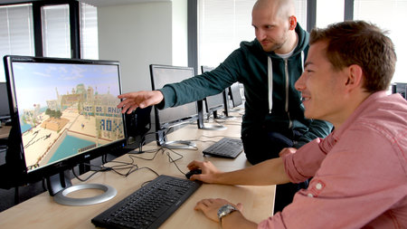 Zwei Männer sitzen an einem Computer und blicken auf einen Bildschirm, auf dem ein Computerspiel läuft.