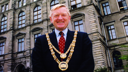 Mann im schwarzen Sacko trägt eine goldene Amtskette und steht vor einem Gebäude.
