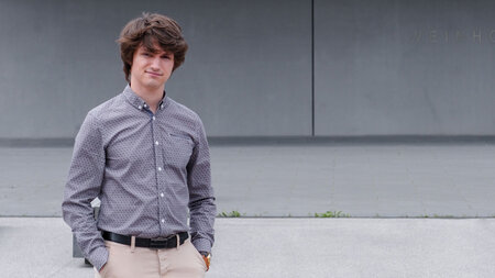 Junger Mann steht mit seinen Händen in den Hosentaschen vor einem grauen Gebäude