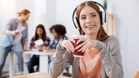 Junge Frau, die Kopfhörer trägt, sitz mit einem Kaffeebecher in der Hand an einem Tisch. Im Hintergrund sprechen drei Personen miteinander.