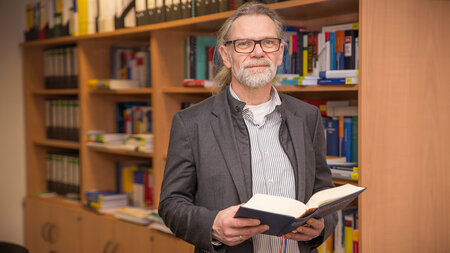 Mann mit Brille steht mit Buch in der Hand vor einem Bücherregal.