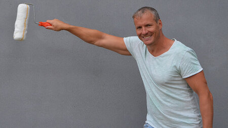 Mann im T-Shirt mit Malerrolle in der Hand steht vor einer grauen Wand.