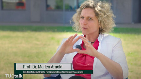 Eine Frau mittleren Alters im Blazer sitzt draußen, spricht und gestikuliert.