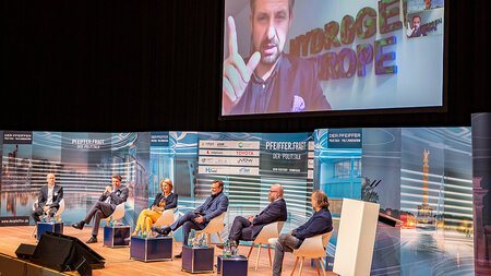 Fünf Männer und eine Frau sitzen auf einem Podium. Ein Mann ist per Konferenz auf einer Leinwand zugeschaltet.