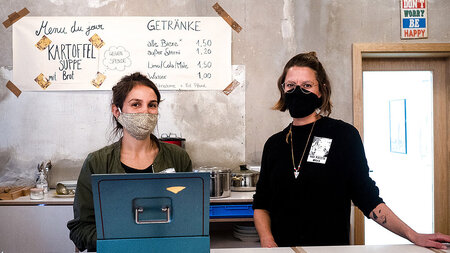 Two young women in masks stand in a restaurant kitchen.
