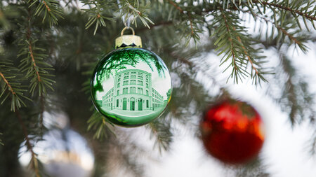 Weihnachtskugeln hängen an einem Zweig eines Nadelbaums, es spiegelt sich ein Gebäude in einer der Kugeln.