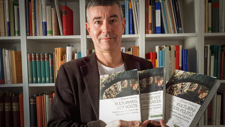 A man in front of a bookshelf holds up three books. One is in German, one is in English, and one is in Czech.