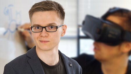 EIn junger Mann im Sakko. Dahinter ein junger Mann mit VR-.Brille.