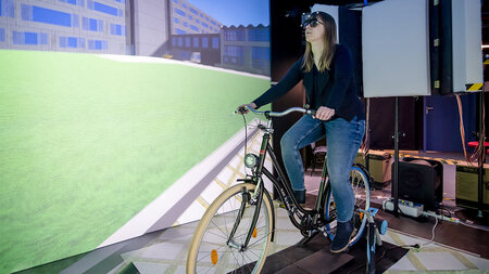 Eine junge Frau sitzt auf einen Fahrrad. Um sie herum ist eine virtuelle Umgebung projiziert.