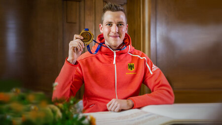 Eine junger Mann sitzt mit einer Medaille an einem Tisch und lacht. Vor ihm liegt ein aufgeschlagenes Buch.