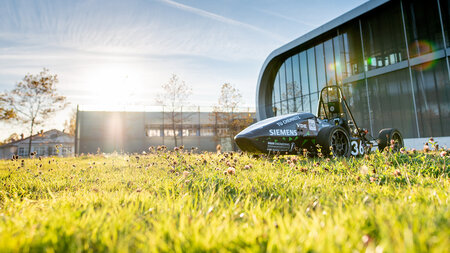 Ein kleiner Rennwagen steht auf dem Rasen vor einem modernen Gebäude, im Hintergrund geht die Sonne unter.