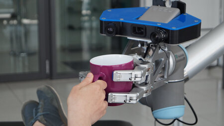 Eine Hand greift nach einer Tasse, die von einem Roboterarm überreicht wird.