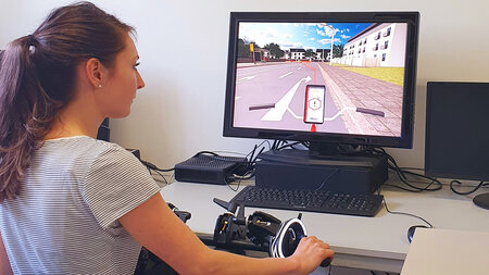 Eine junge Frau sitzt an einem Computer und nutzt einen Fahrraadsimulator. 