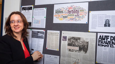 Eine Frau mittleren Alters mit schulterlangen Haaren steht an einer Posterwand.