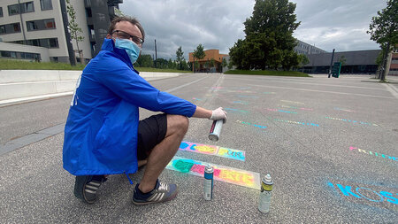 Ein junger Mann in Regenjacke trägt eine OP-Maske und hält eine Spraydose.