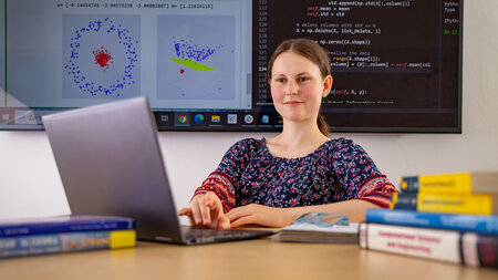 Junge Frau arbeitet an einem Laptop, im Hintergrund ist der Ausschnitt einer Darstellung auf einem großen Bildschirm zu sehen.