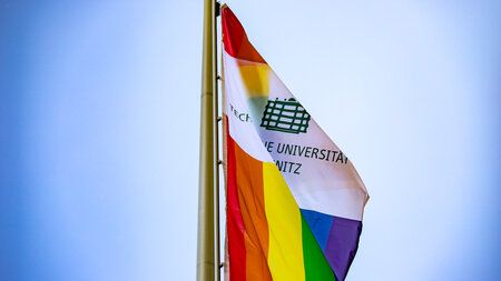 Drei Regenbogenflaggen mit dem Logo der TU Chemnitz flattern im Wind.