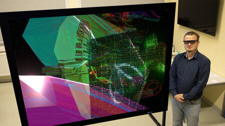 Ein junger Mann mit VR-Brille steht neben einem Bildschirm. 