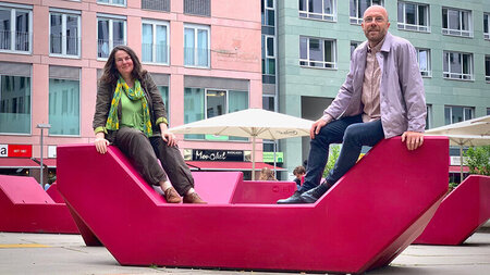 Eine Frau und ein Mann sitzen im Freien auf einer roten Bank.