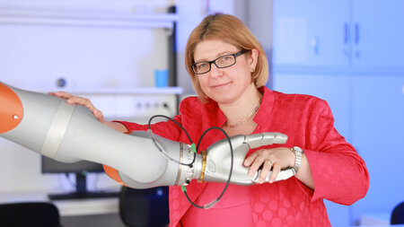Eine Frau mittleren Alters mit schulterlangen Haaren und Brille gibt einem Roboter die Hand.