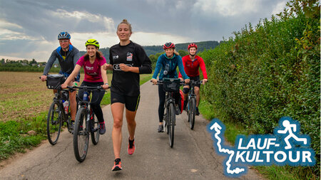 Eine Läuferin und vier Radfahrer bewegen sich auf einem Radweg durch eine flache Landschaft.