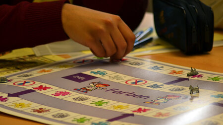 Eine Hand schiebt eine Spielfigur über ein farbiges Spielfeld.