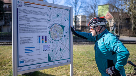 Ein Radfahrer mit einem Helm steht vor einem Informationsschild, das auf einer Landkarte eine Route zeigt. 