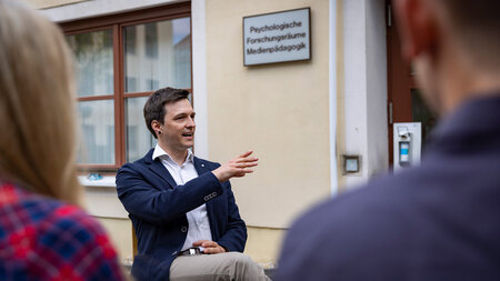 Ein junger Mann im Jacket sitzt auf einer Bank und spricht mit zwei Personen.