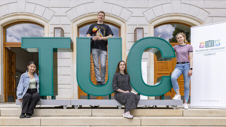 Drei junge Frauen und ein junger Mann stehen an der Buchstabenfolge TUC.