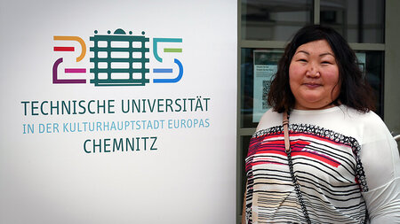 Eine ältere asiatische Frau steht neben einem Aufsteller mit dem Logo der TU Chemnitz.