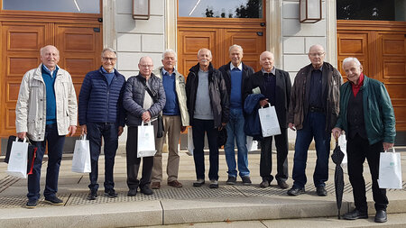 Neun ältere Männer stehen vor dem Eingang zu einem historischen Gebäude.