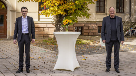 Zwei Männer im Anzug stehen links und rechts von einem Tisch, auf dem ein Strauß Blumen steht.