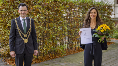 Ein Mann im Anzug und mit goldener Amtskette steht neben einer jungen Frau mit Urkunde und Blumenstrauß in den Händen.