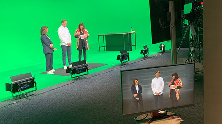 Ein Mannn und zwei Frauen stehen in einem Studio vor einer grünen Wand und werden beim Diskutieren gefilmt. Das Bild wird auf einen Monitor im Studio übertragen.