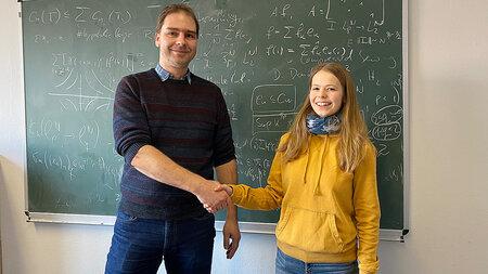 A young man and a girl stand in front of a blackboard and shake hands.