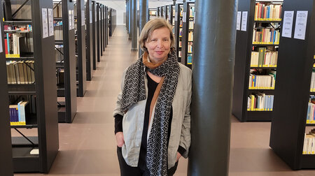 Eine Frau lehnt in einer Bibliothek, in der viele mit Büchern gefüllte Regale stehen, an einer schwarzen Säule.