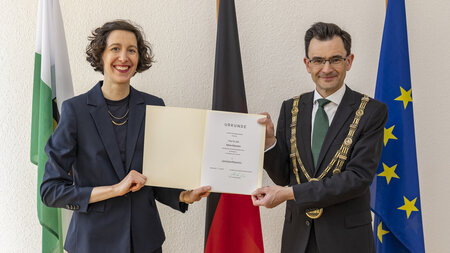 Eine Frau im Kleid und mit schulterlangen Haaren und ein Mann im Anzug mit großer Kette halten beide eine Urkunde. 