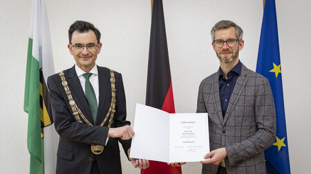 Ein Mann im Anzug und mit einer großen Kette und ein Mann im Sakko mit Brille halten gemeinsam eine Urkunde.