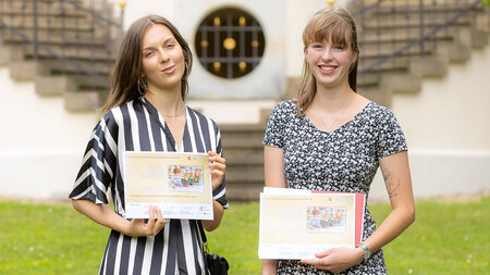 Zwei junge Frauen stehen nebeneinander und halten jeweils eine Urkunde in den Händen. 