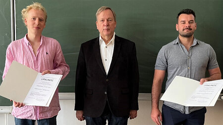 Three men stand side by side in front of a blackboard, two holding certificates in their hands.