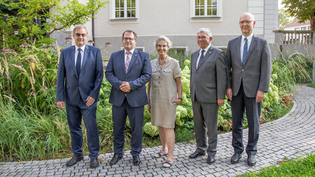 Vier Männer und eine Frau in formeller Kleidung stehen vor einem Gebäude und lachen bzw. lächeln. 