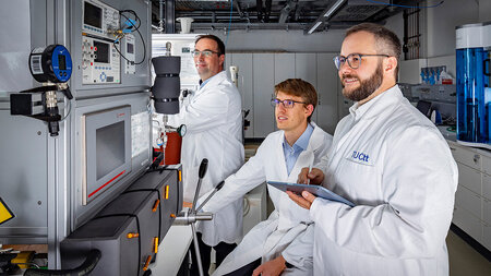 Three men in lab suites stand in front of a machine.