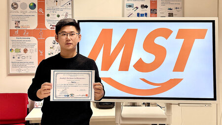 Ein junger asiatischer Mann mit Brille hält ein Urkunde in den Händen. Hinter ist mit dem Buchstaben MST die Abkürzung der Professur Mess- und Sensortechnik zu sehen.
