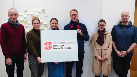 Sechs Personen stehen nebeneinander und halten eine Tafel mit einem Logo hoch.