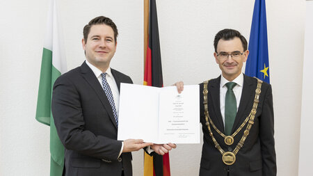 Zwei Männer im Anzug stehen nebeneinander, lächeln und halten eine Urkunde.